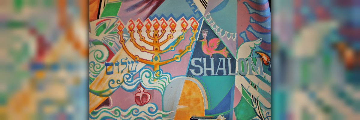 Ein Ausschnitt des Wandbehangs der jüdischen Kultusgemeinde. Zu sehen sind ein Chanukkaleuchter und das Wort Shalom vor buntem Hintergrund. 