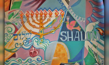 Ein Ausschnitt des Wandbehangs der jüdischen Kultusgemeinde. Zu sehen sind ein Chanukkaleuchter und das Wort Shalom vor buntem Hintergrund. 