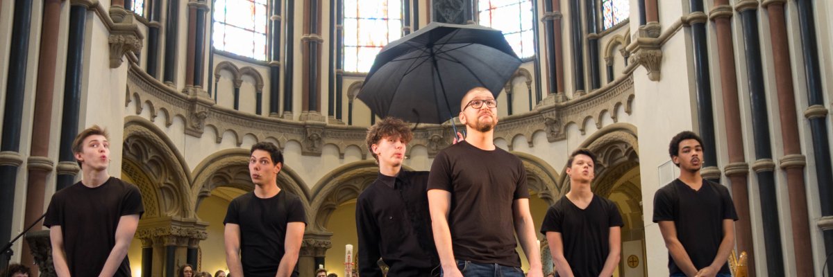 Aschermittwoch_Koblenz_Jugend Im Vordergrund stehen zwei Männer. Der eine hält einen offenen Regenschirm über den anderen. Im Hintergrund stehen vier weitere Männer, welche ihre geschlossenen Stabregenschirme vor sich auf den Boden stellen. Dies geschieht vor dem Altar der Herz-Jesu-Kirche.